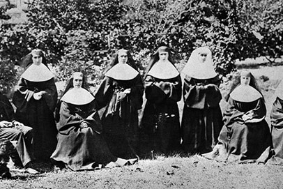 A black and white historical photo of eight nuns dressed in habits and a priest wearing a top hat posing in a garden.
