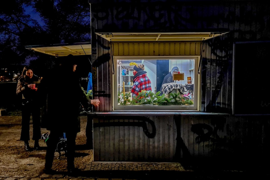 People stand with steaming cups at night outside of a store made out of corrugated iron.