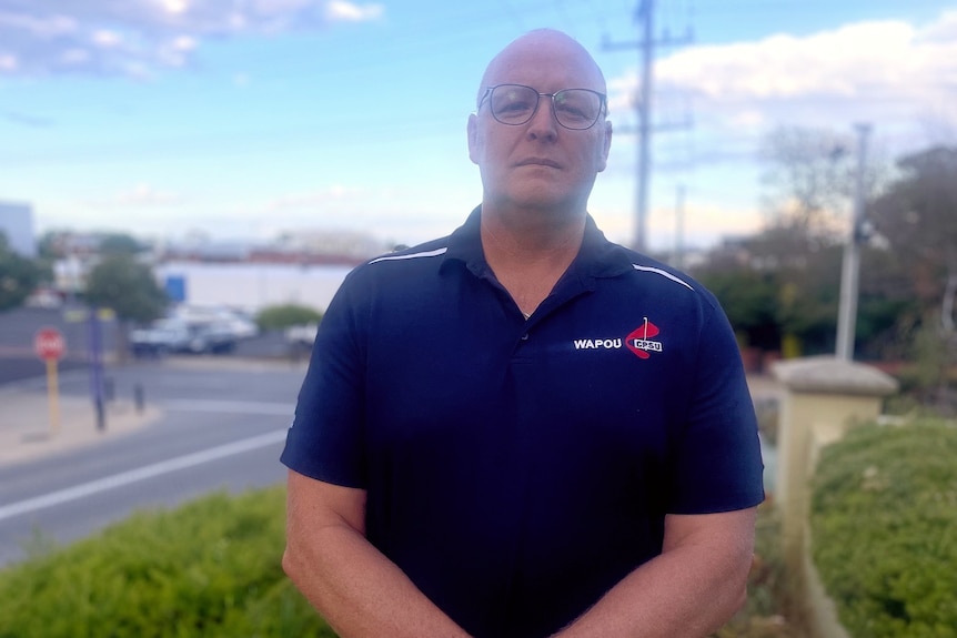 A bespectacled, middle-aged man with a shaved head stands on a suburban street, looking solemn.