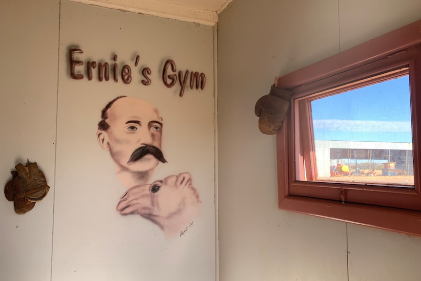 A painting on a wall says 'Ernie's Gym', balding man, handlebar moustache, a camel. Blue skies visible from window. 