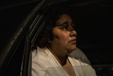 Ebony wears a white shirt and sits in the back of her car, image taken through window