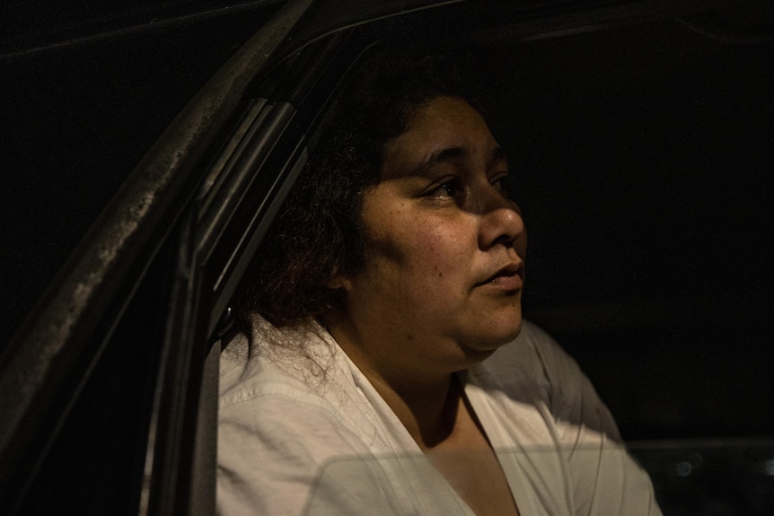 Ebony wears a white shirt and sits in the back of her car, image taken through window