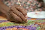 A seated person's hand holds a kebab stick to create a dot shape on colourful dotted painting on canvas.