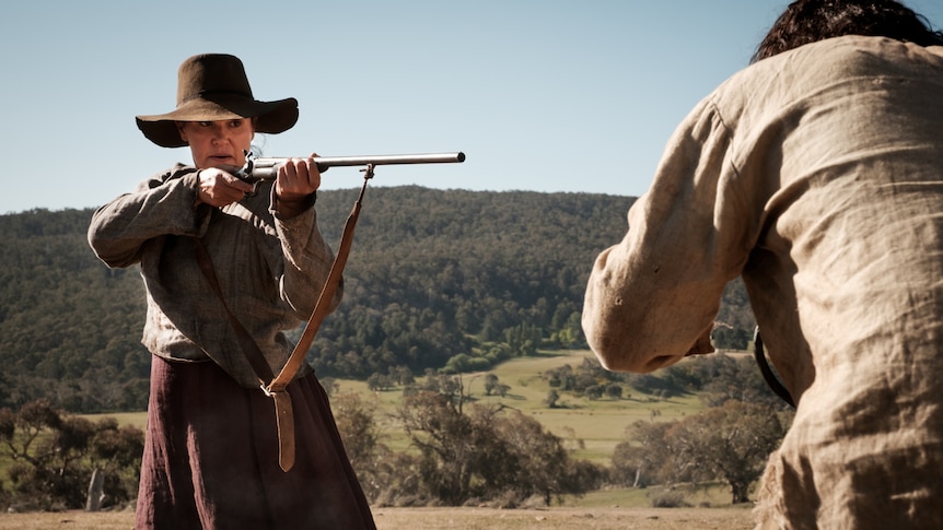 An Aboriginal woman in a wide-brimmed hat and colonial dress point a rifle at a cowering man