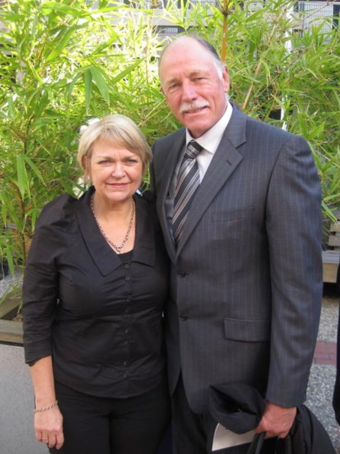 A woman with blonde hair wearing a black long-sleeved, collared shirt stands smiling next to a man in a grey pinstriped suit.
