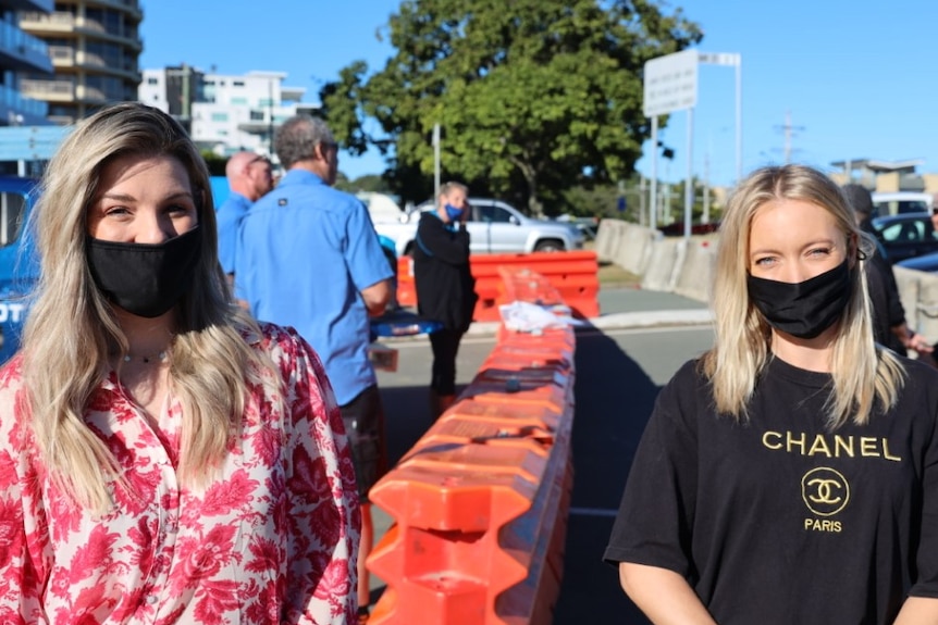 Two women wearing face masks stand either side of barricade with blue sky behind them