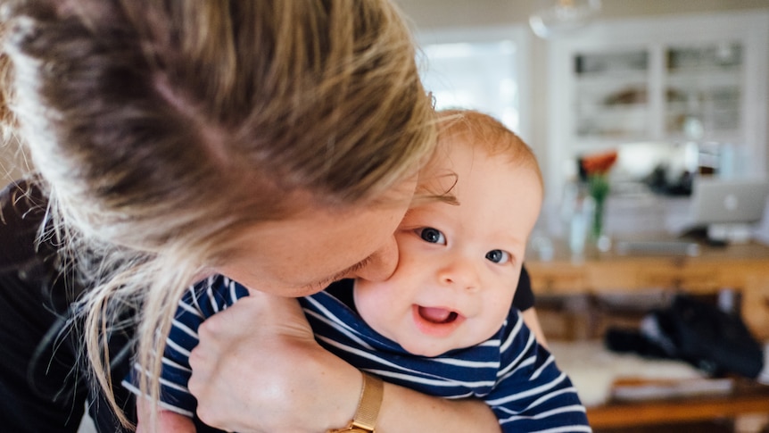 A close up shot of the top of a woman's head nestled into a small baby, who looks out smiling widely.