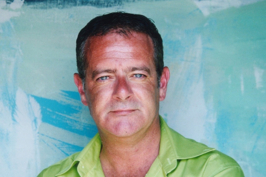 A man in a green shirt looks at the camera with a blue wall behind him.