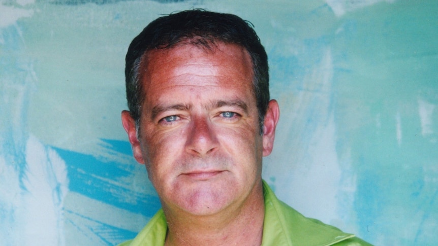 A man in a green shirt looks at the camera with a blue wall behind him.