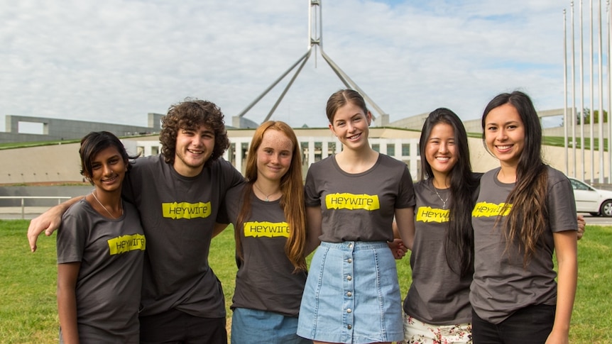 a group of six young people in matching black t-shirts pose in front of parliament house in canberra