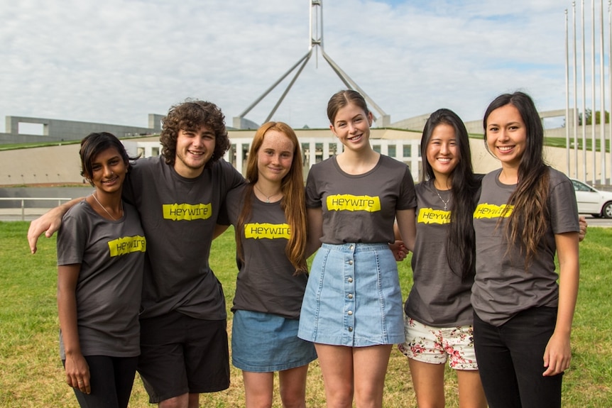 a group of six young people in matching black t-shirts pose in front of parliament house in canberra