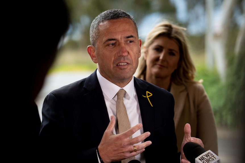 A man with short hair wearing a dark suit and gold tie with a blonde woman behind him