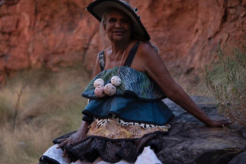 A woman wearing an elaborate dress and peacock-feathered hat in front of red rocks.