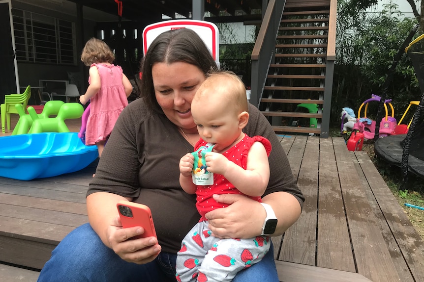 Dana holds a phone while Lucy sits in her lap watching and eating a yoghurt.