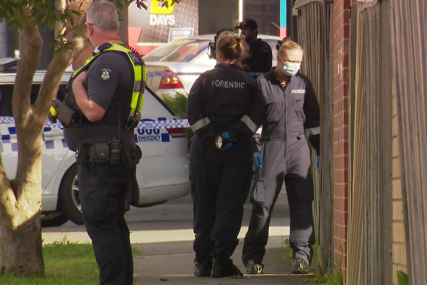 A male police officer looks towards a police car while two female forensic investigators enter a property.
