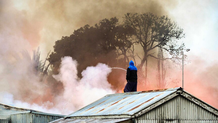 A man stands on the roof of a shack and squirts a garden hose on what is obviously a large fire, out of sight.
