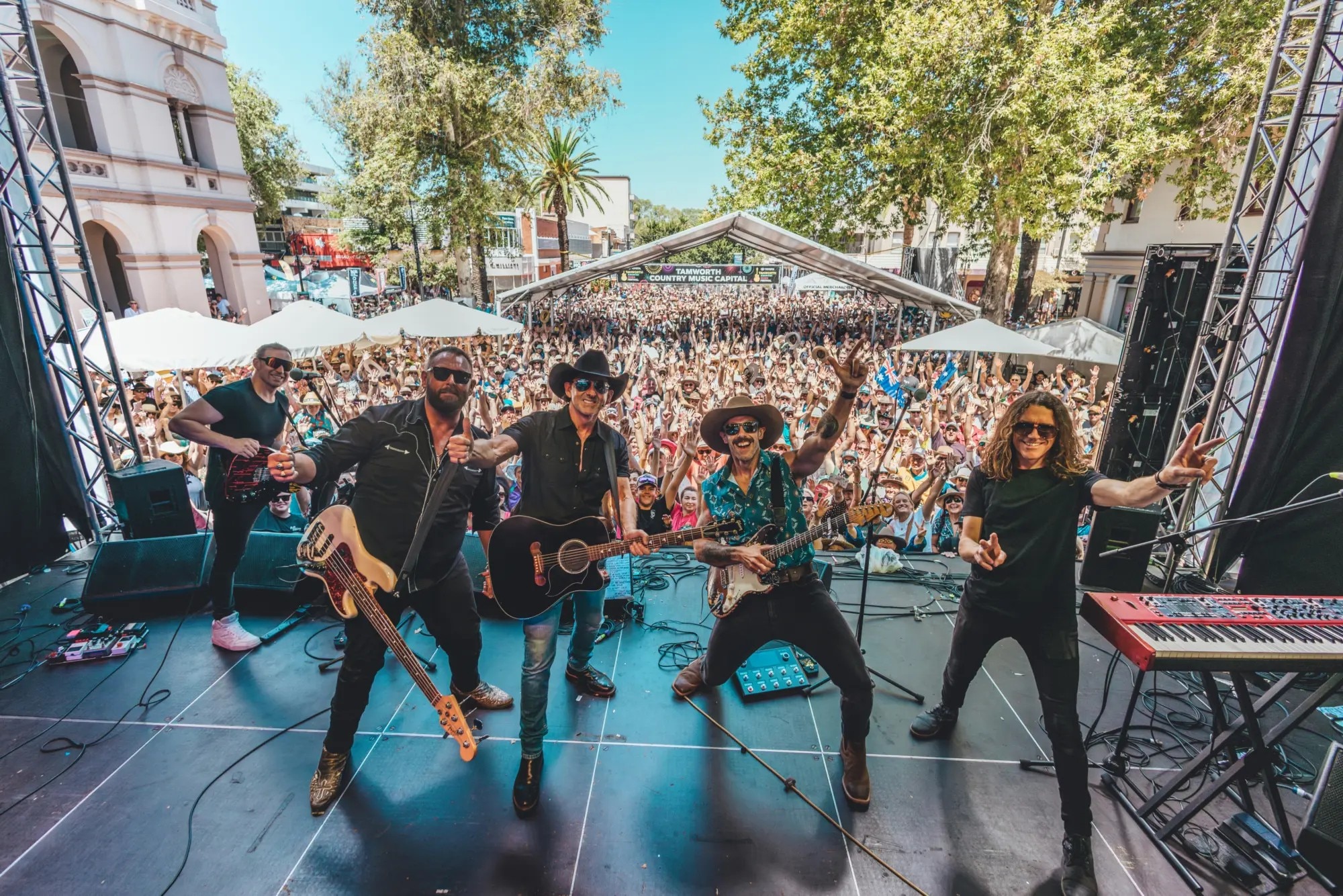 Uma banda está no palco posando para a câmera, atrás dela uma multidão aplaude