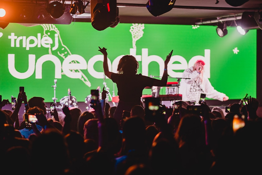 As Tones & I performs on stage in front of a green triple j Unearthed digital banner, someone in the crowd sits on shoulders.