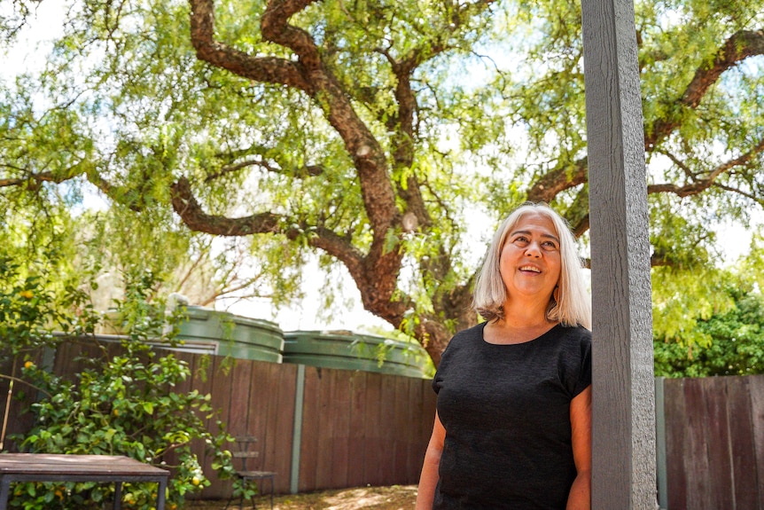 A woman wearing a black t-shirt leans against a wooden pole in a leafy backyard.