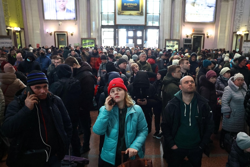 A large crowd of people, some using mobile phones, gather inside a building a look towards information boards.