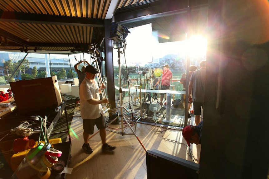 Production crew setting up lights and cameras on balcony overlooking sporting precinct with sun in shot.