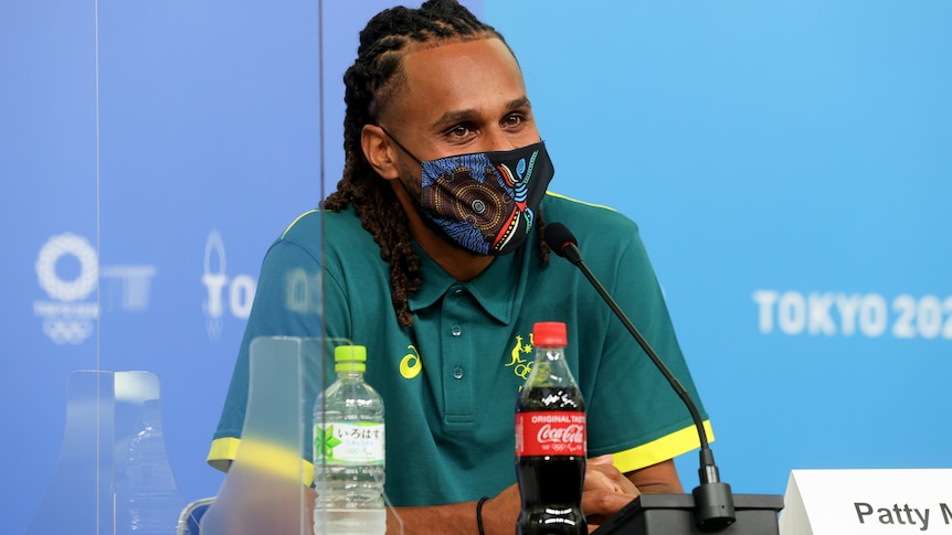 Wearing a mask with Indigenous art printed on it, Patty Mills sits in front of a microphone