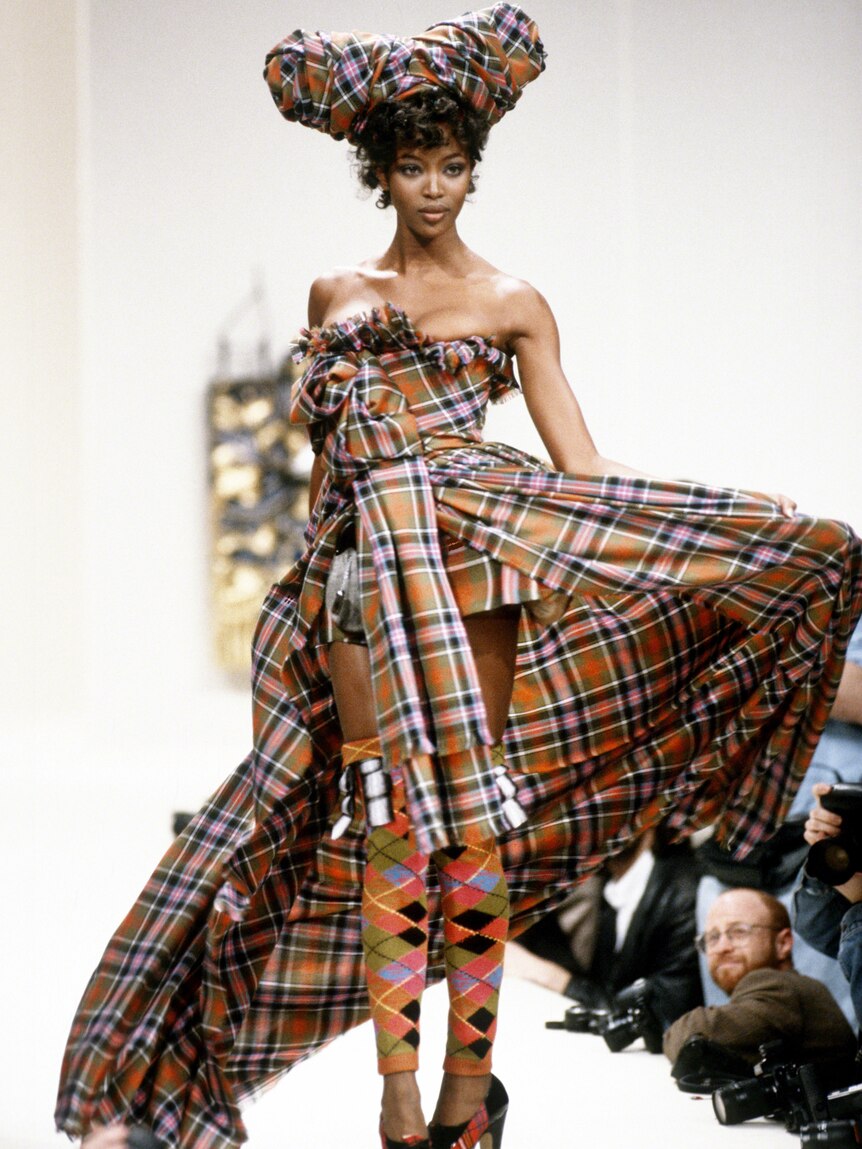Noami Campbell in a dress and headwear walks down a fashion runway