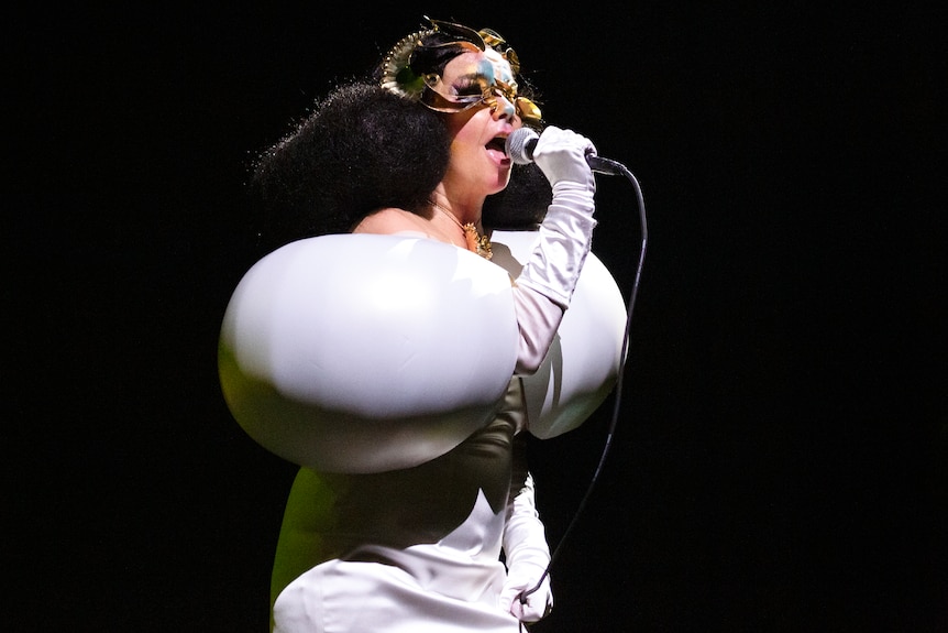 Bjork sings into a microphone on stage. She wears a white outfit with hugely inflated sleeves and a lot of eye makeup