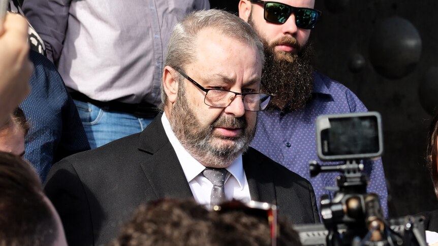 A man, Brett Button, standing in a crowd of people with news cameras surrounding him