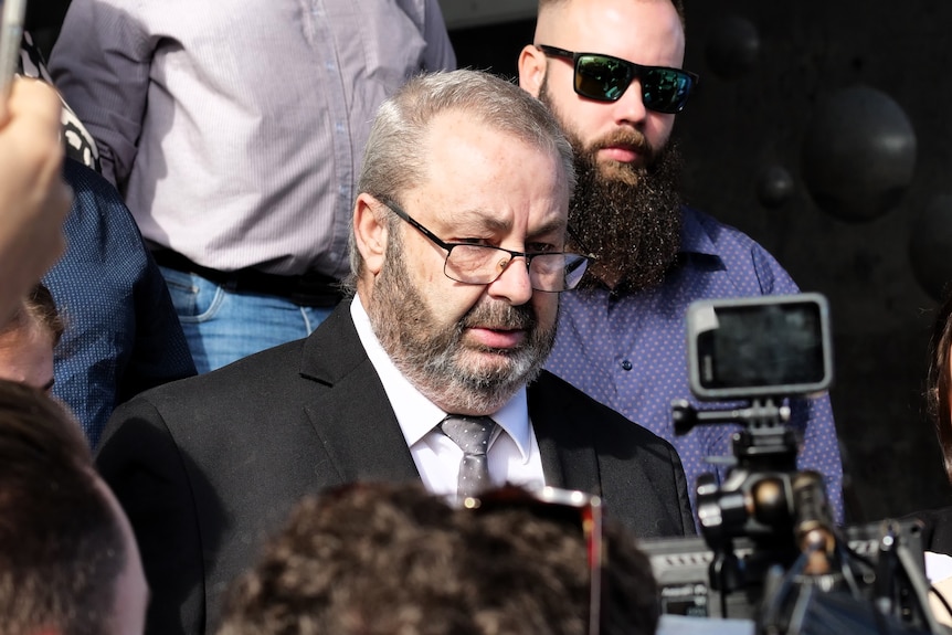 A man in a dark suit stands outside, surrounded by journalists.