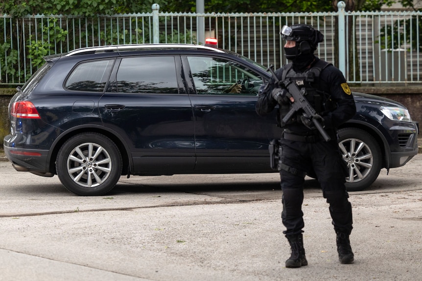 A masked man in dark combat clothes holding a rifle stands in front of a dark car.