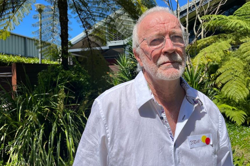 An older man with white hair and a white beard standing in a leafy courtyard on a sunny day.