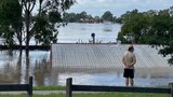 A man stands on a footpath in front of flood water that is up to the tin roof of a shed 