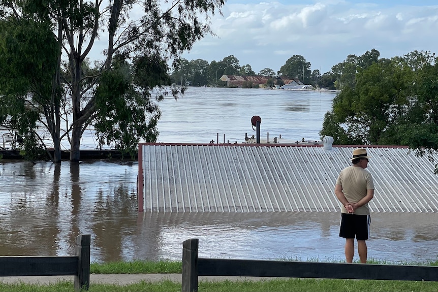 A man stands on a footpath in front of flood water that is up to the tin roof of a shed 