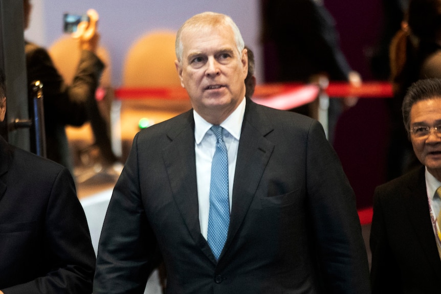 Prince Andrew in a black suit with blue tie walks