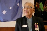 An old Chinese woman wearing a jacket with WW2 medals stands in front of an Australian flag.