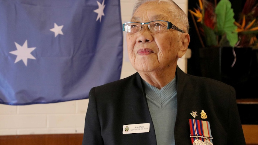 An old Chinese woman wearing a jacket with WW2 medals stands in front of an Australian flag.