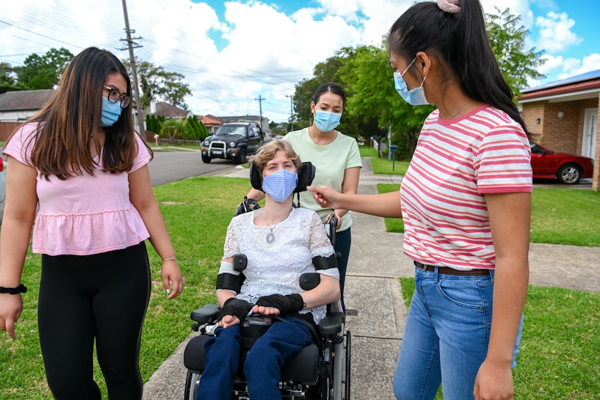 Rachel in her wheelchair on the footpath with three carers, all in masks.