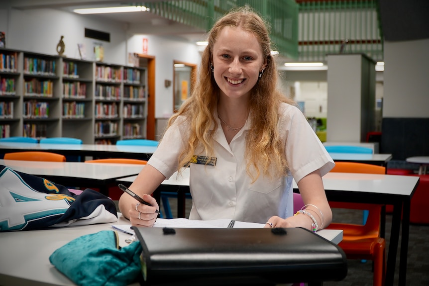 Teenage girl in school uniform smiles happily as she sits at desk in school library with study notes in front of her.