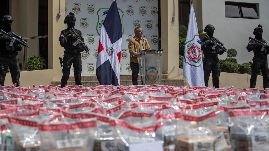Heavily armed men flank a man speaking at a podium in front of thousands of plastic bags and flanked by Dominican flags.