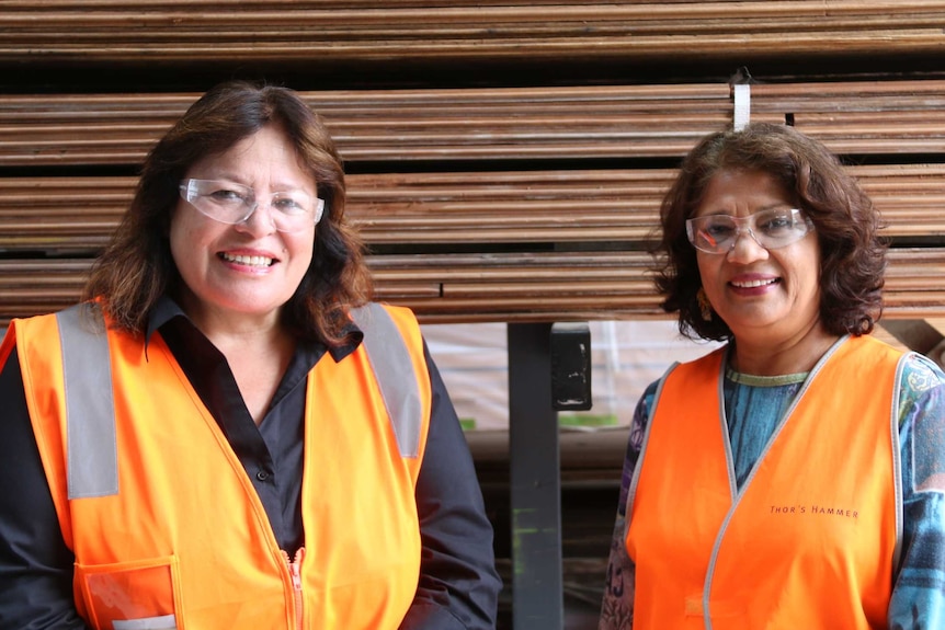 Two women in high-vis vests and safety glasses stand in front of planks of wood.