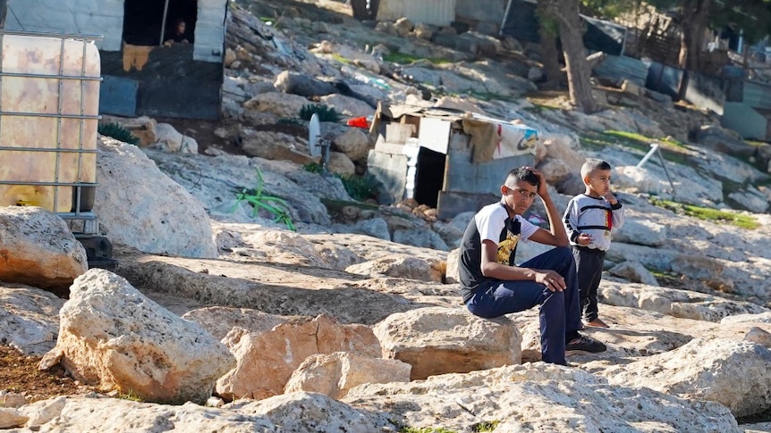 Two young children sitting on a rock, surrounded by structures made of scrap metal