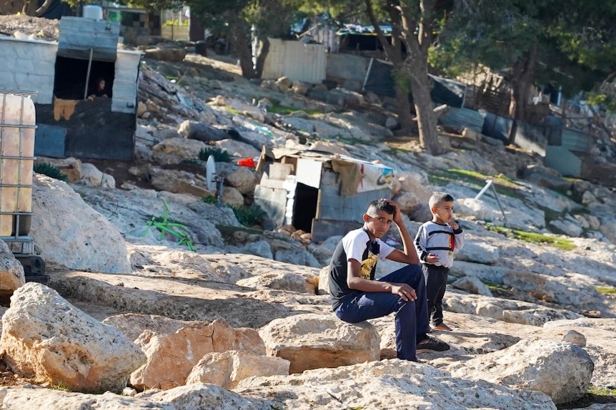 Two young children sitting on a rock, surrounded by structures made of scrap metal