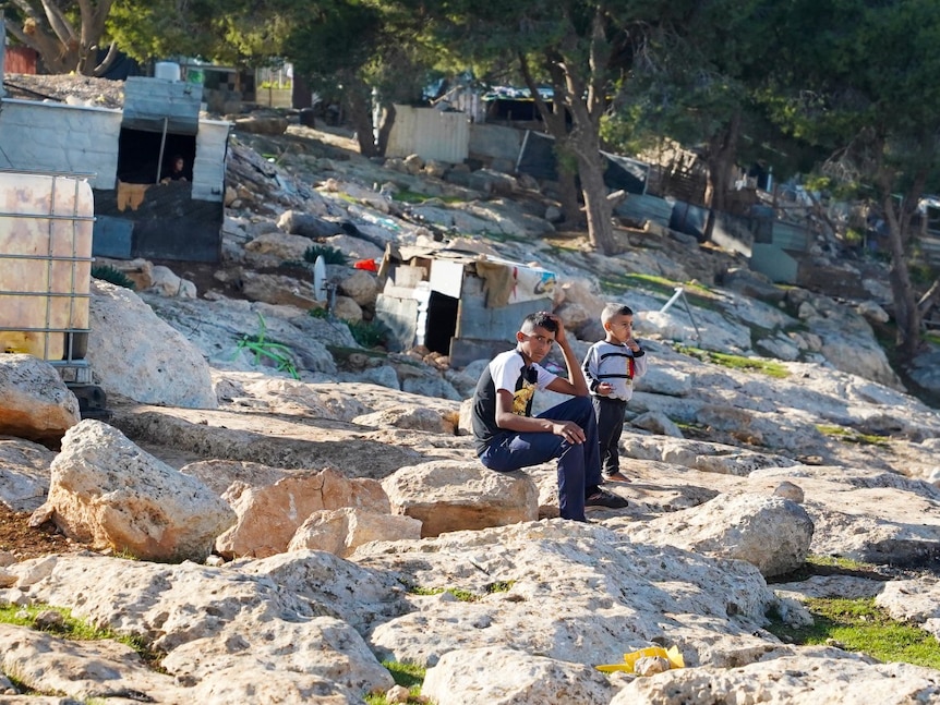 Two young children sitting on a rock, surrounded by structures made of scrap metal