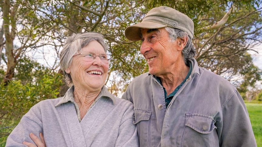 An older woman stands smiling at on old man in a cap next to her, trees behind them.