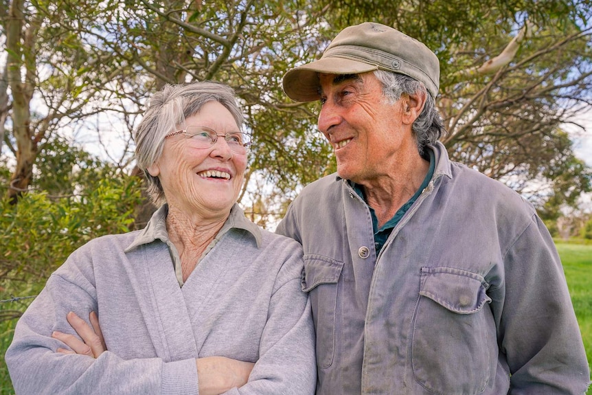 An older woman stands smiling at on old man in a cap next to her, trees behind them.