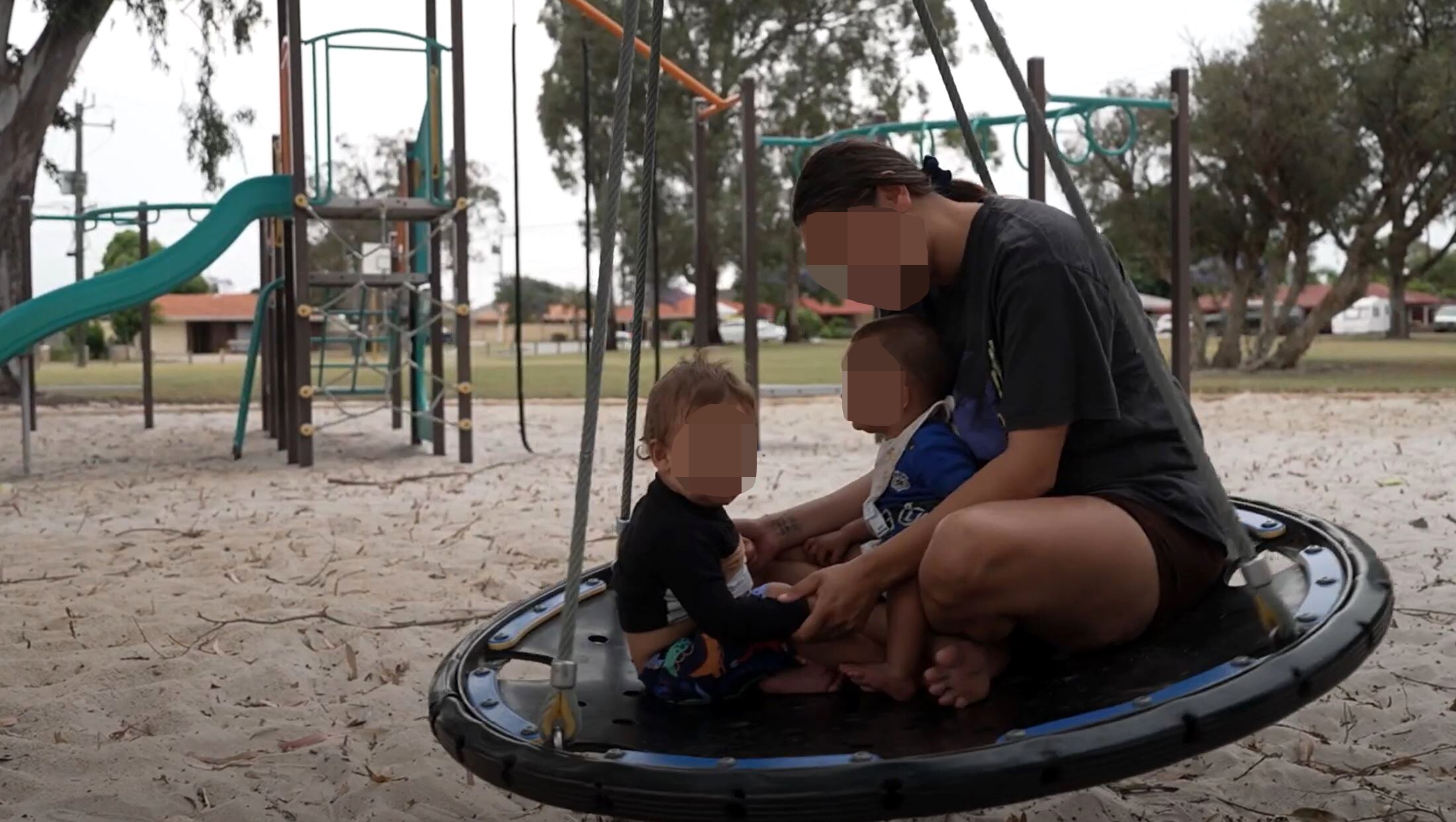 An unidentified mother and two young children on a round swing in a playground.