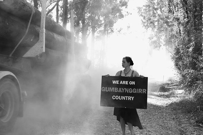 An indigenous woman stands in a forest holding a sign which says 'we are on Gumbaynggirr country'.