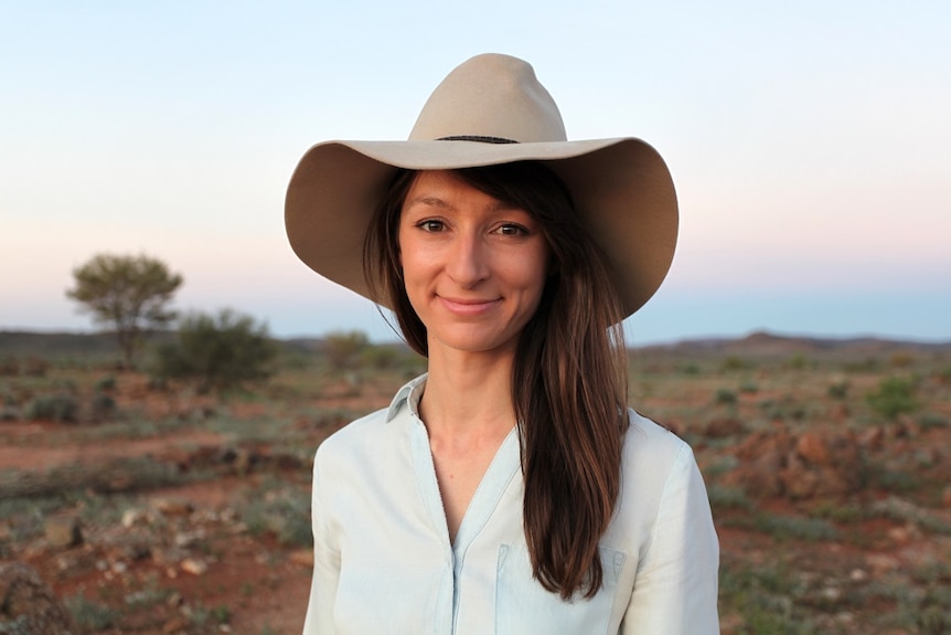 a smiling woman in a collared short and akubra wide-brimmed hat stands before a wide landscape of scrubby bush and red dirt