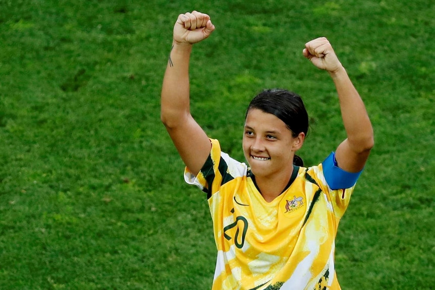 Sam Kerr raises her arms in a strong-woman stance, looking to the crowd with a smile.